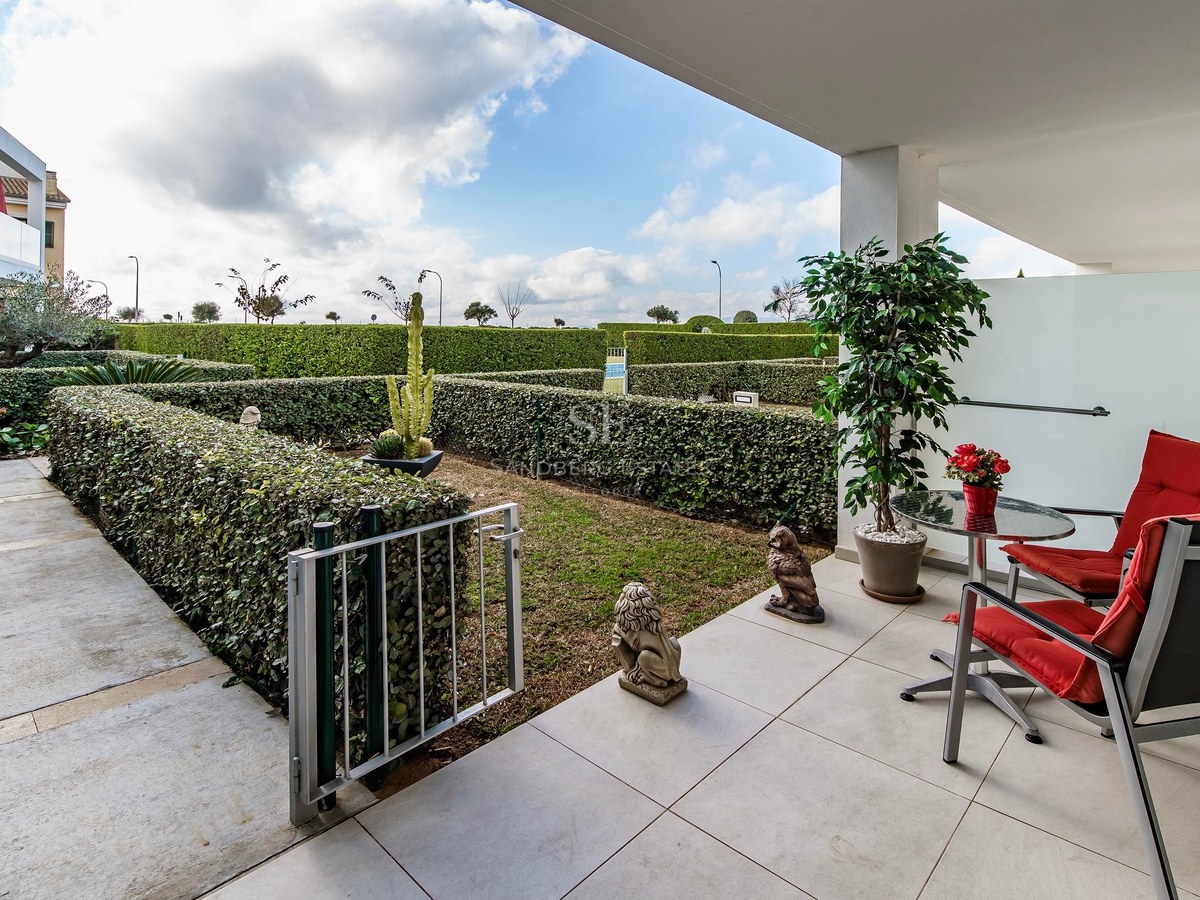 Terrasse blanche moderne avec deux chaises rouges et une table donnant sur un jardin privé aux haies taillées.