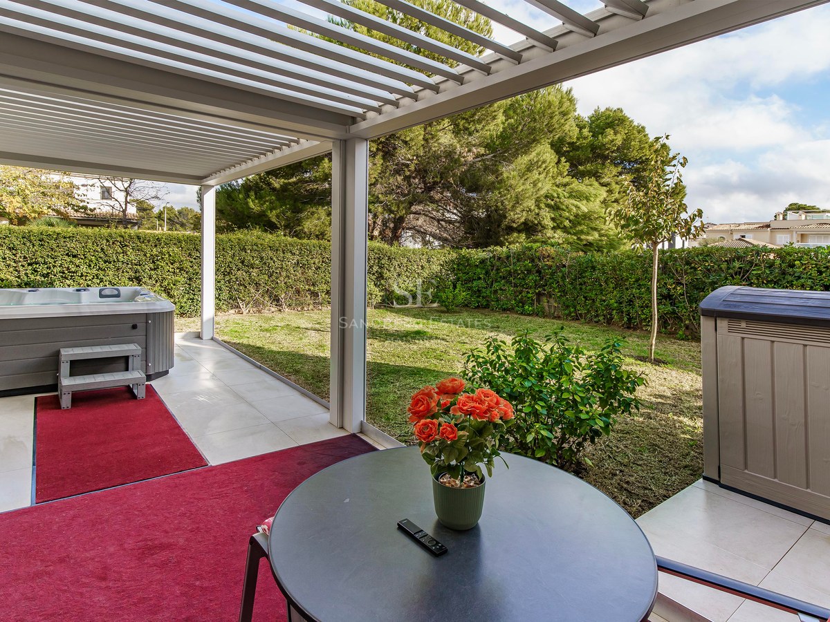 Terrasse avec pergola blanche, jacuzzi gris, tapis rouges et vue sur un jardin verdoyant avec haies.