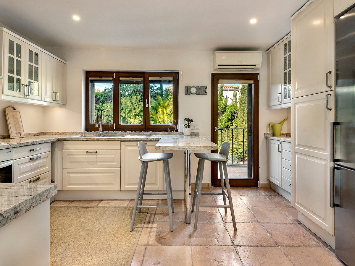 Cocina luminosa con armarios crema, encimeras de granito, suelos de terracota y vistas a la exuberante vegetación.
