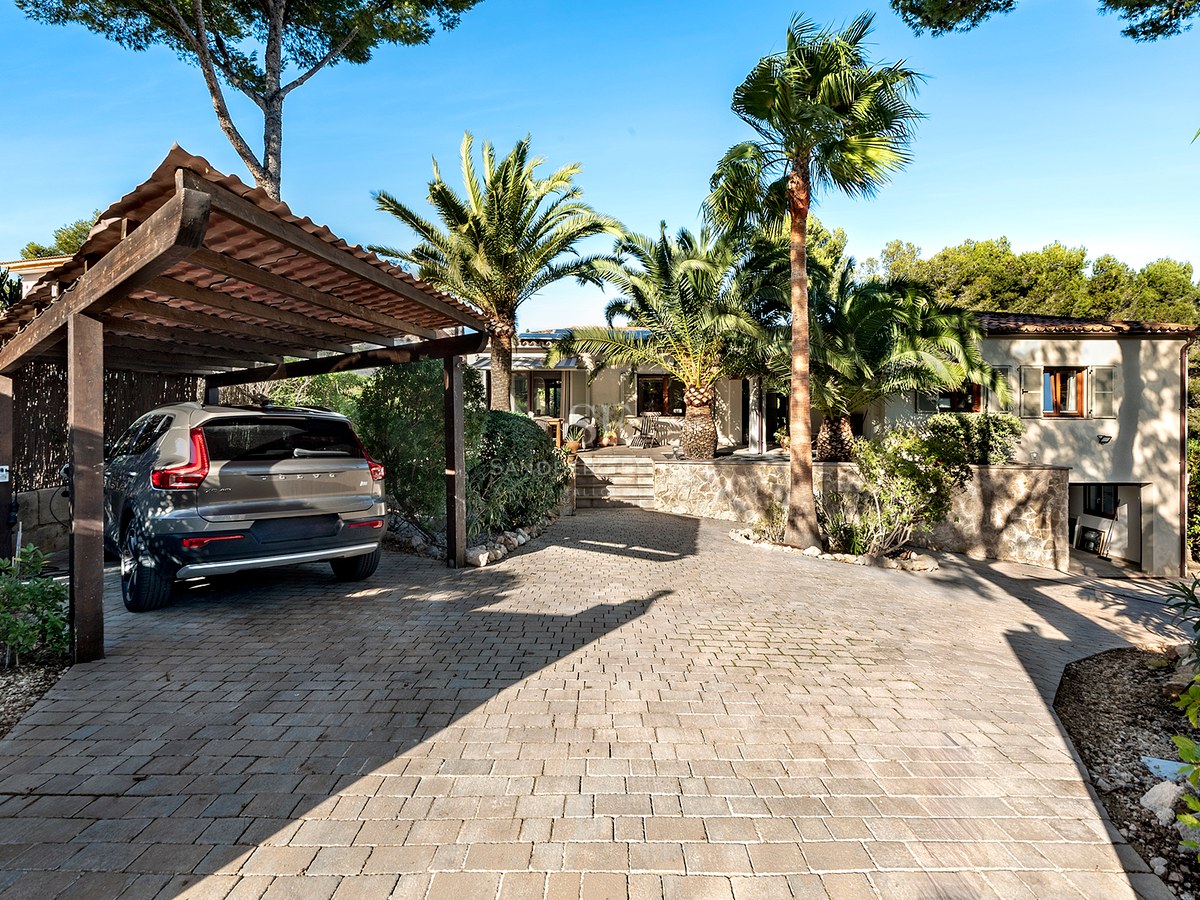 Camino empedrado con porche de madera para coche, palmeras y fachada de villa al fondo bajo un cielo despejado.