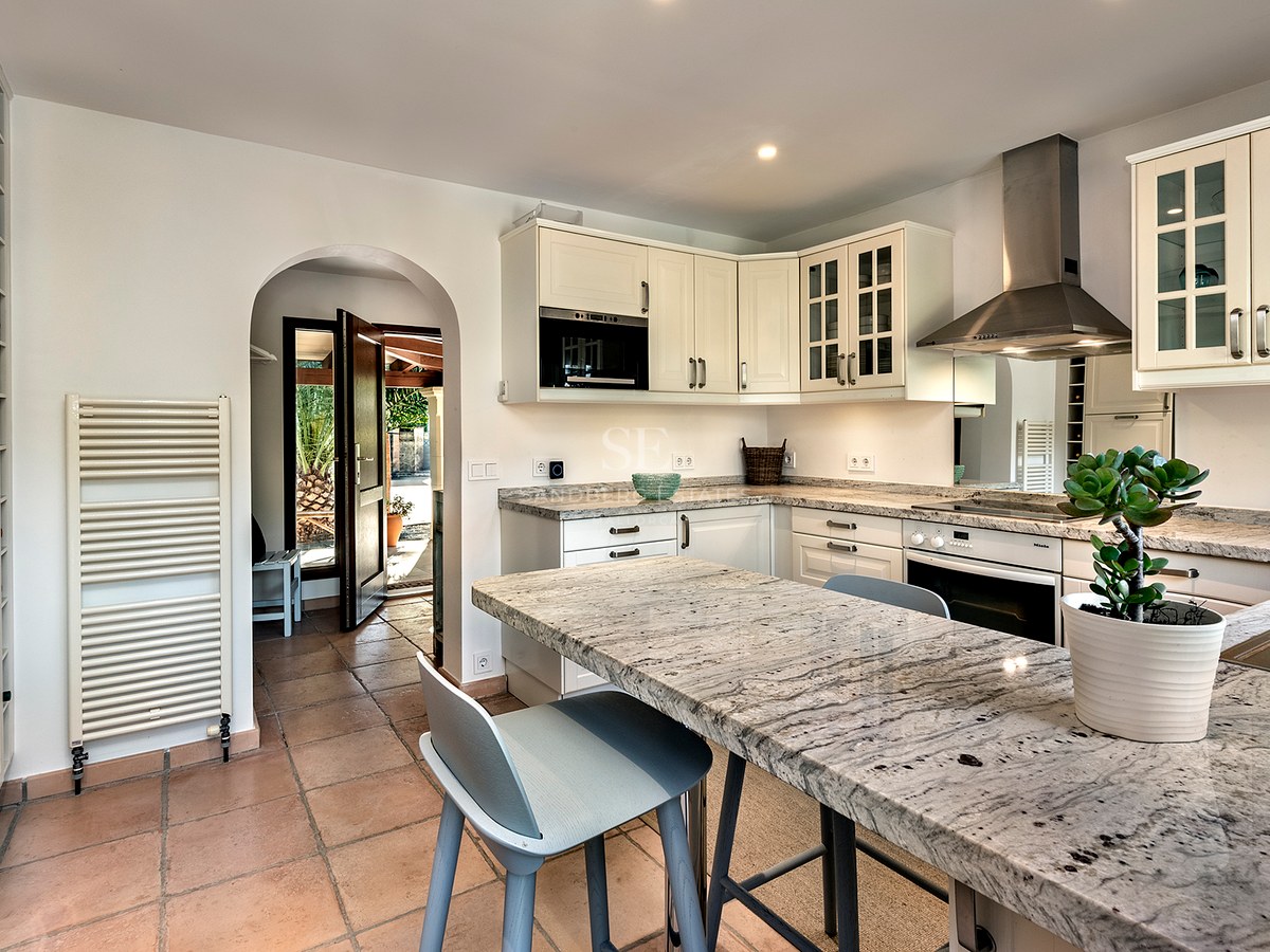 Cocina luminosa con armarios blancos, encimeras de granito, suelos de terracota y un arco que da al exterior.