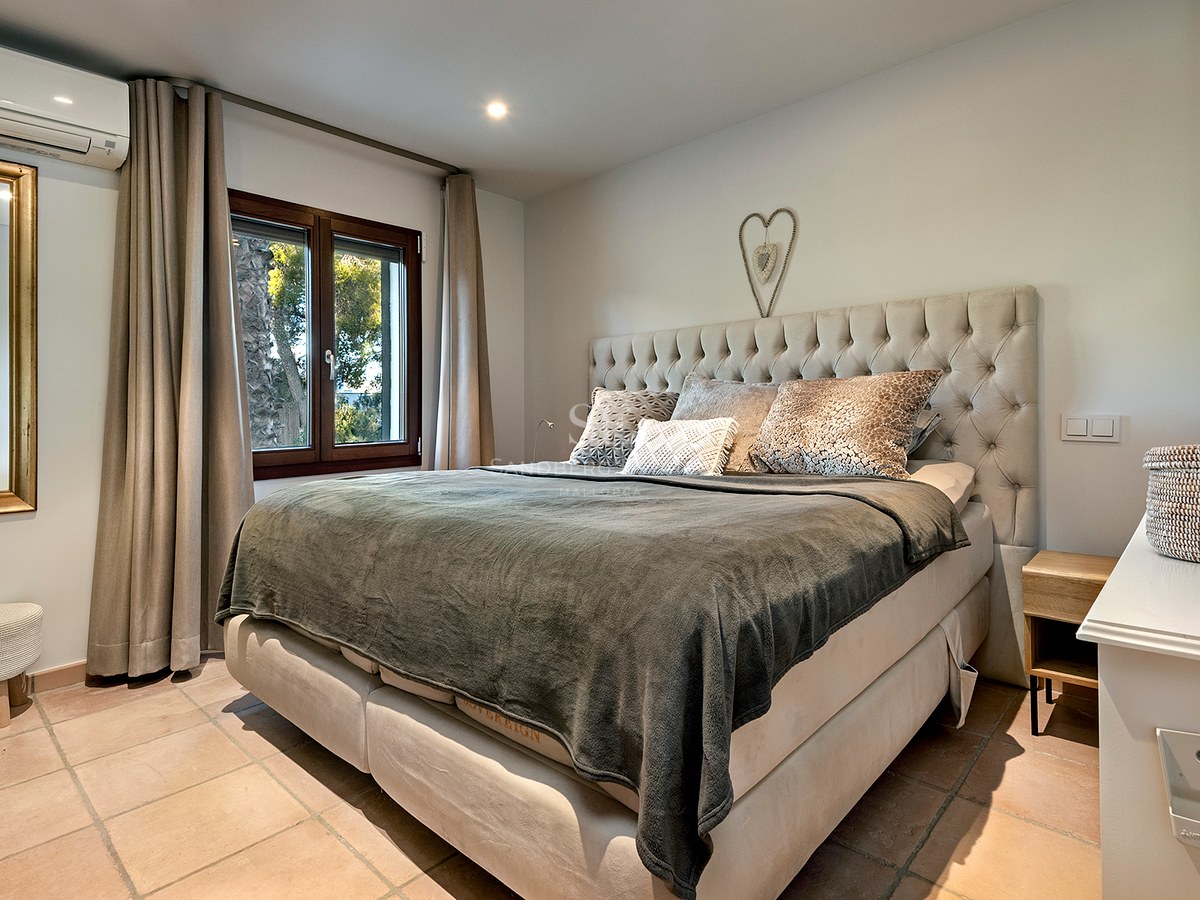 Dormitorio acogedor con una cama grande, cabecero capitoné beige, suelos de terracota y ventana con vistas al jardín.