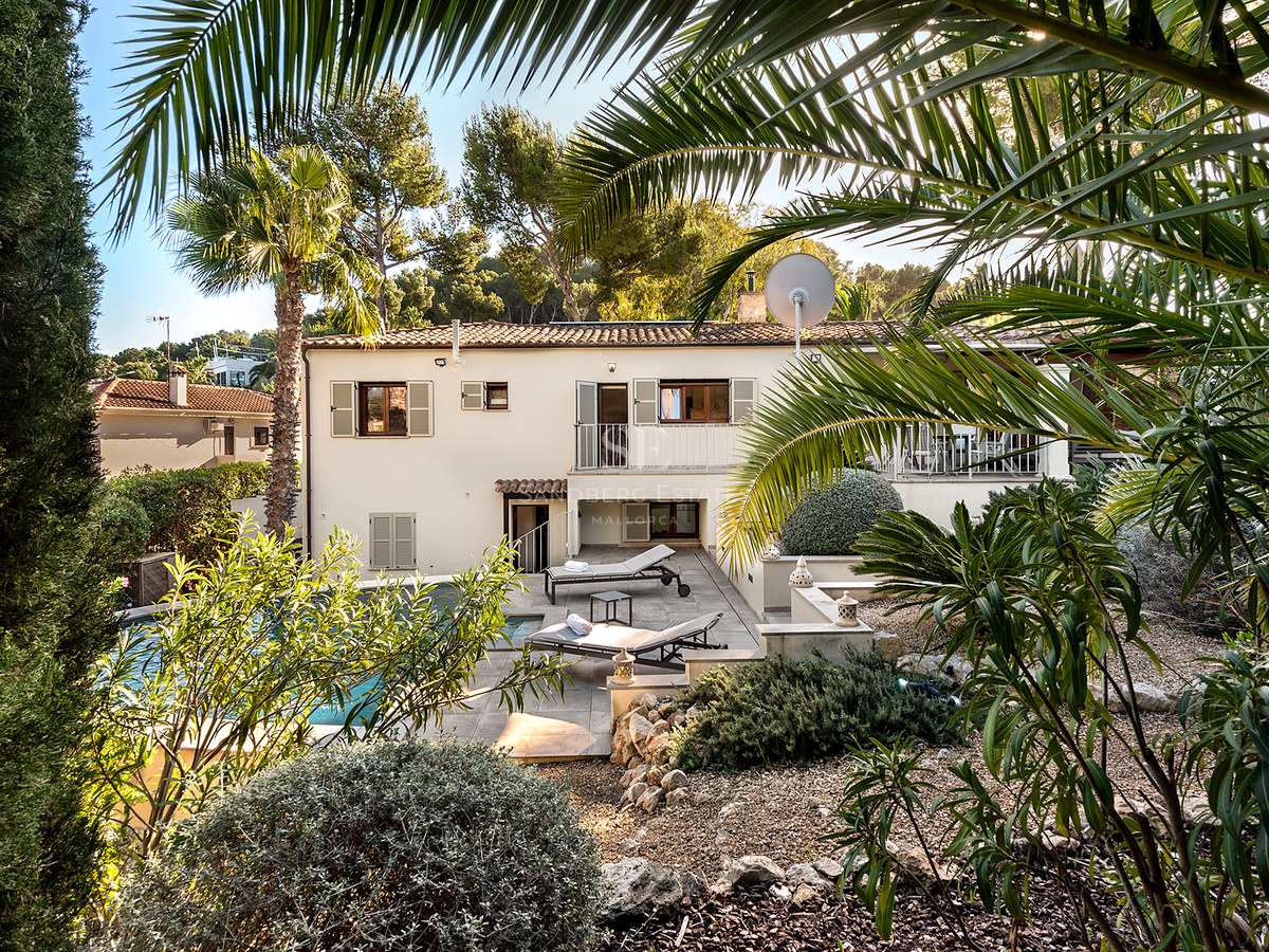 Villa de dos plantas con piscina privada, tumbonas y exuberante vegetación mediterránea bajo un cielo despejado.