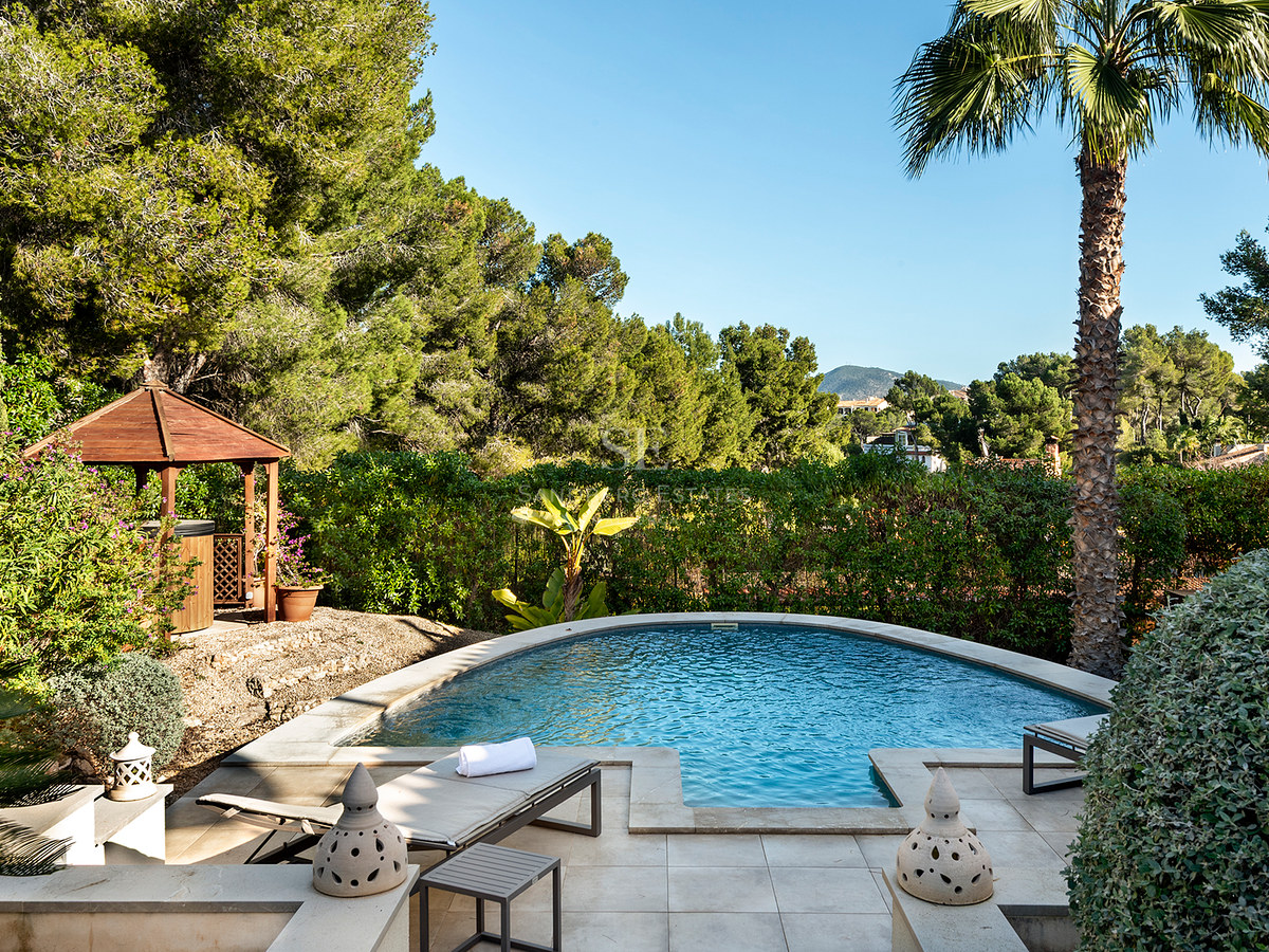 Una piscina curva rodeada por una terraza de piedra, tumbonas, un cenador de madera y vegetación mediterránea densa.