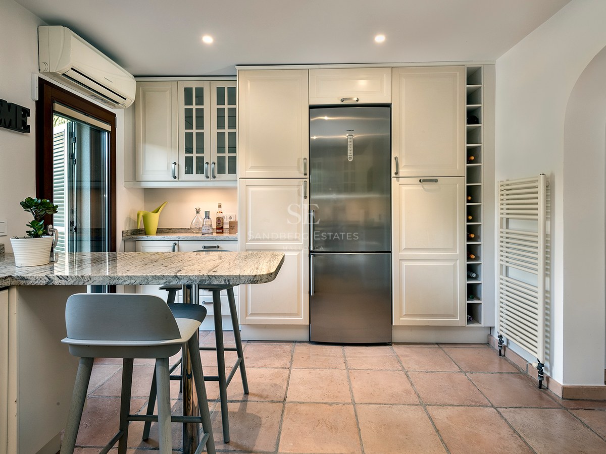 Cocina luminosa con armarios crema, frigorífico de acero inoxidable, suelo de terracota y barra de granito con taburetes.