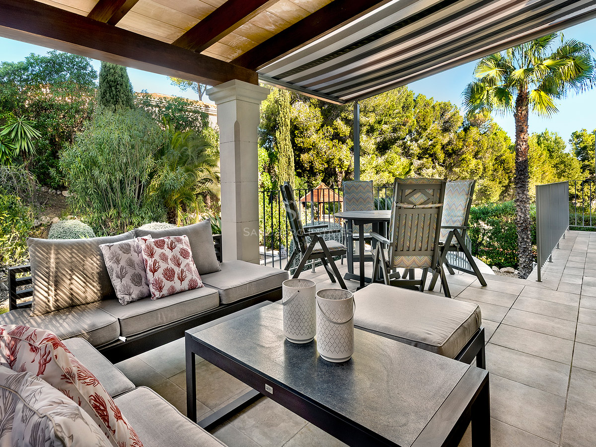 Terraza exterior cubierta con sofás, mesa de comedor y vistas a un frondoso jardín mediterráneo.