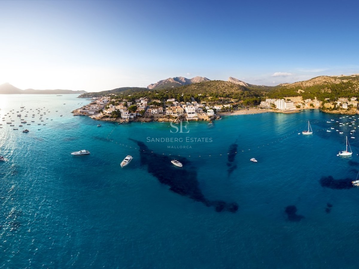Panorama aéreo de una bahía mediterránea con aguas turquesas, barcos blancos y un pueblo costero frente a las montañas.