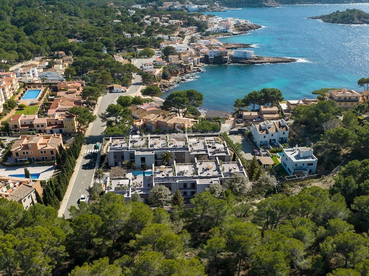 Vista de dron de un complejo de apartamentos moderno entre bosques de pinos y una bahía mediterránea turquesa.