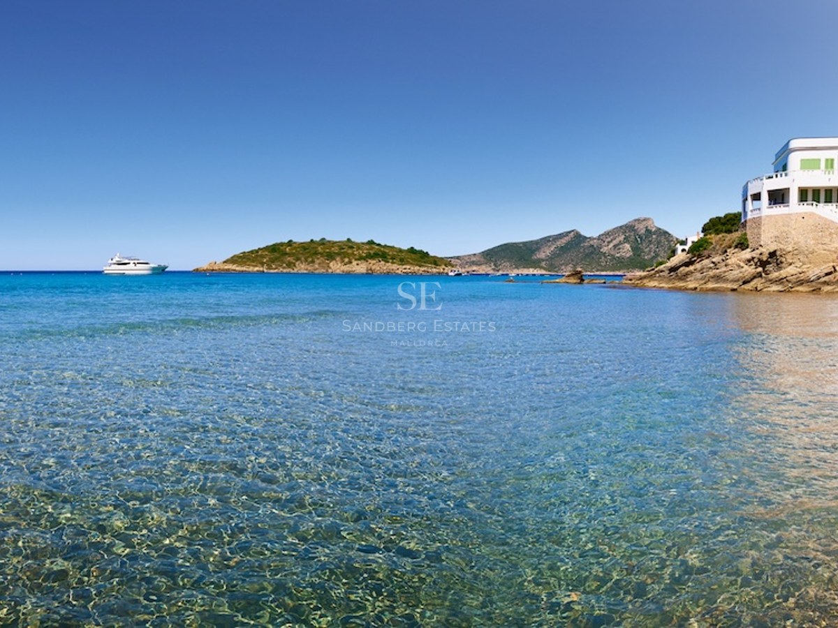 Vista de una bahía de aguas cristalinas turquesas con una villa blanca en un acantilado y montañas al fondo.