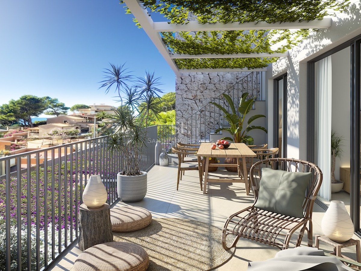 Terraza moderna y soleada con mesa de comedor de madera, sillón de mimbre y pérgola con plantas y vistas a la costa.