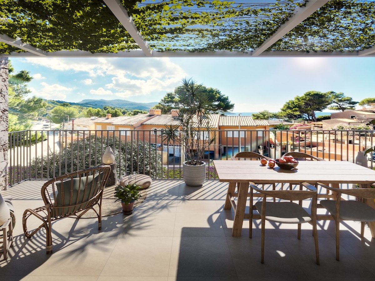 Terraza soleada con mesa de madera, silla de mimbre y pérgola con vides con vistas al paisaje mediterráneo.