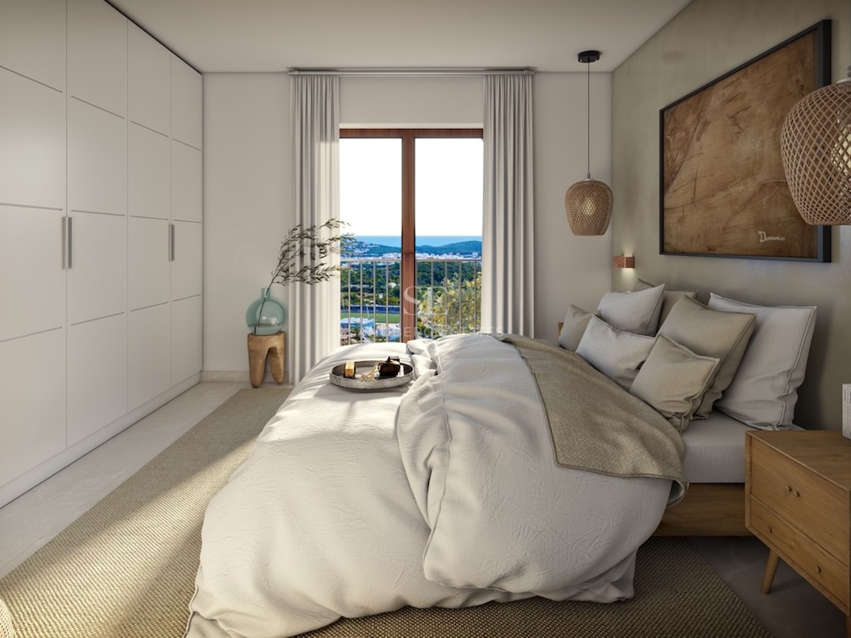 Elegant master bedroom with white linens, built-in wardrobes, and a balcony overlooking a coastal landscape.