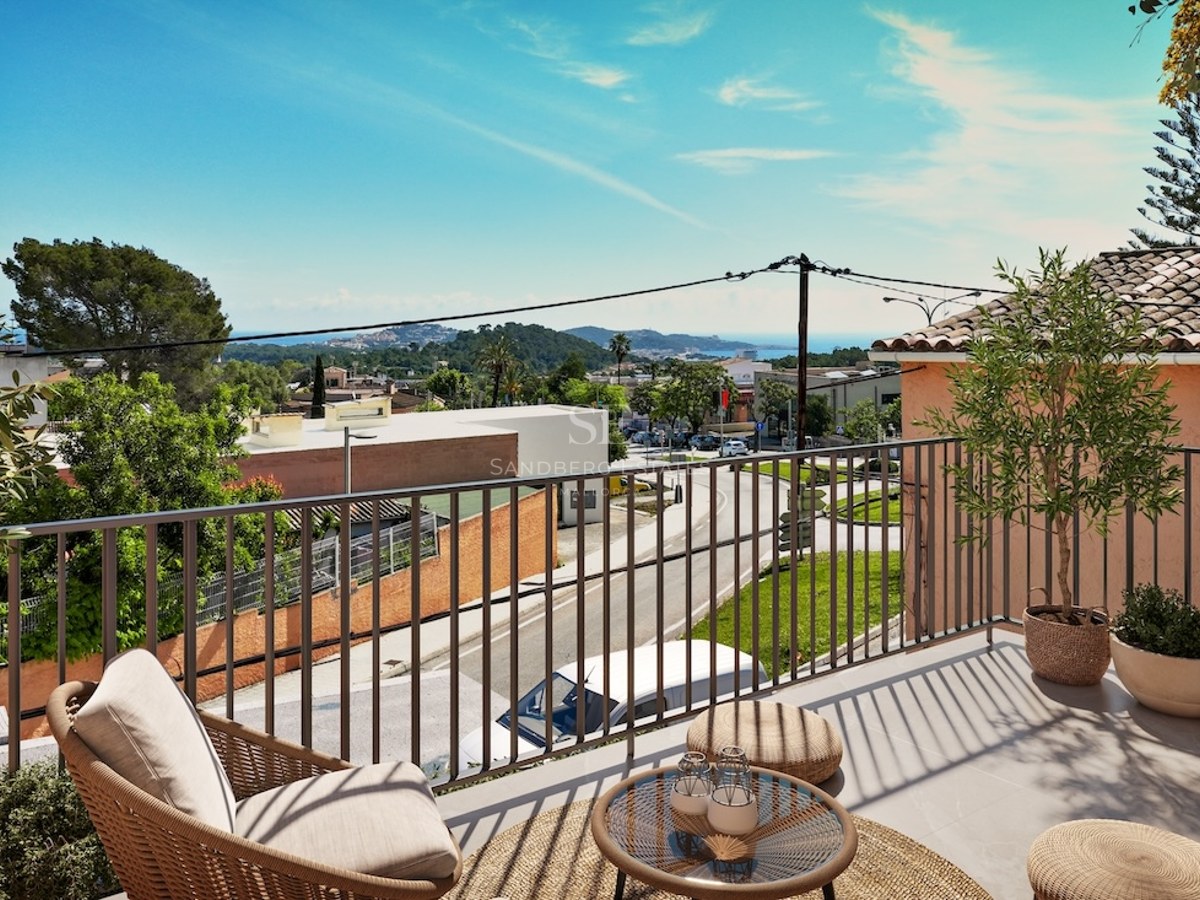 A sunny elevated terrace with a wicker armchair and small table overlooking a coastal town and the sea.