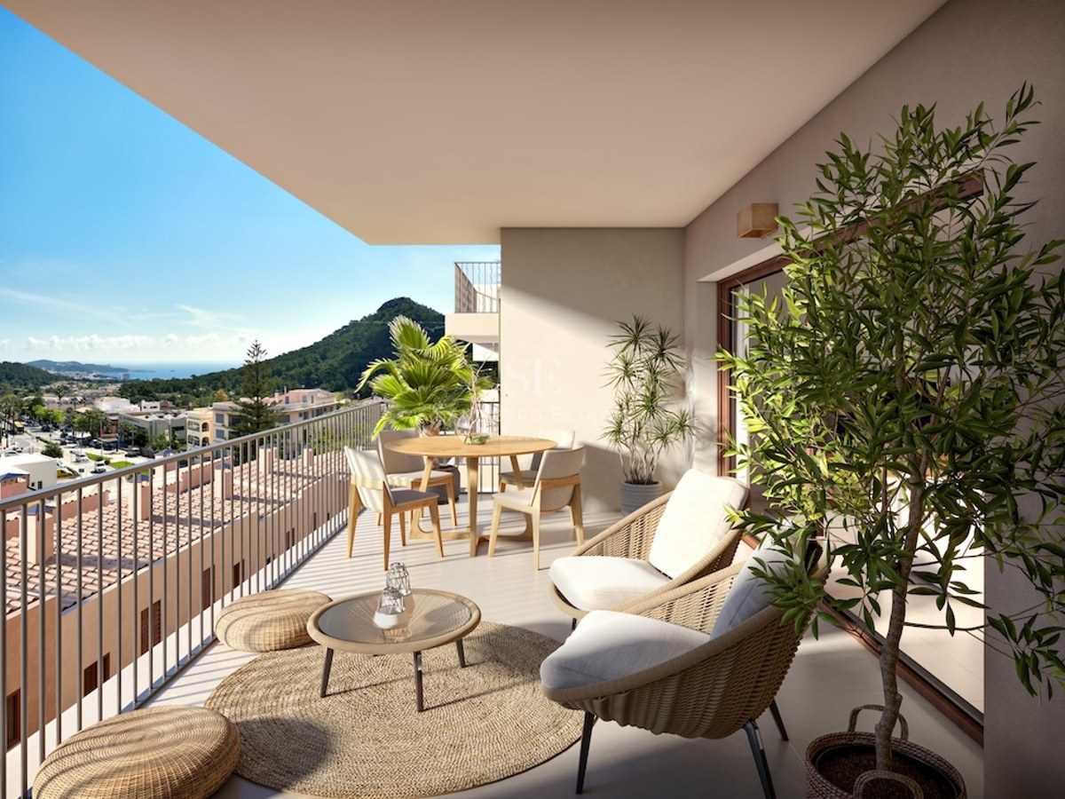 Balcón moderno con muebles de mimbre, mesa de comedor y plantas con vistas a un pueblo costero y colinas verdes.