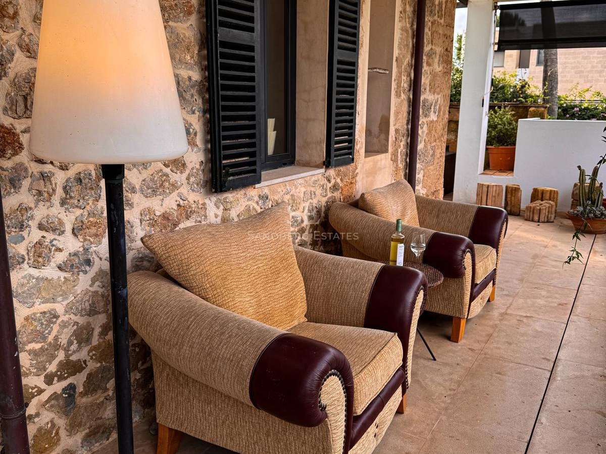 Zwei gepolsterte Sessel und eine Stehlampe auf einer schattigen Terrasse mit mallorquinischer Steinwand und Fensterläden.