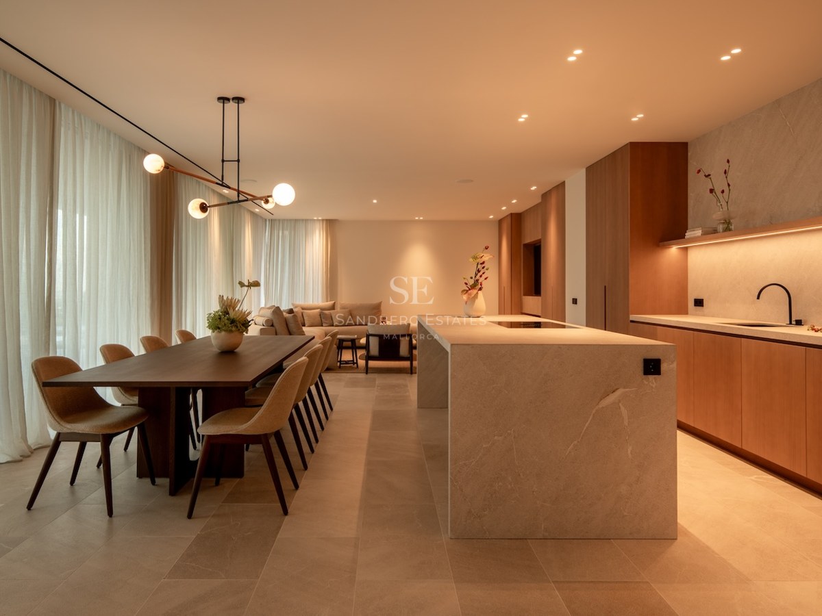 Warmly lit open-plan room with a stone kitchen island, dining table, and sofa behind soft curtains.