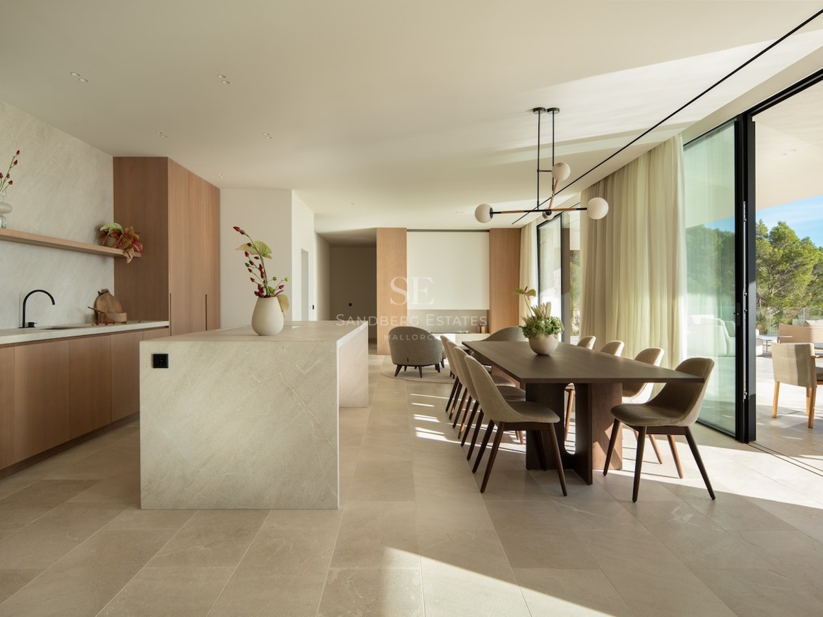 Modern dining room with a wooden table, stone kitchen island, and large floor-to-ceiling windows overlooking a forest.