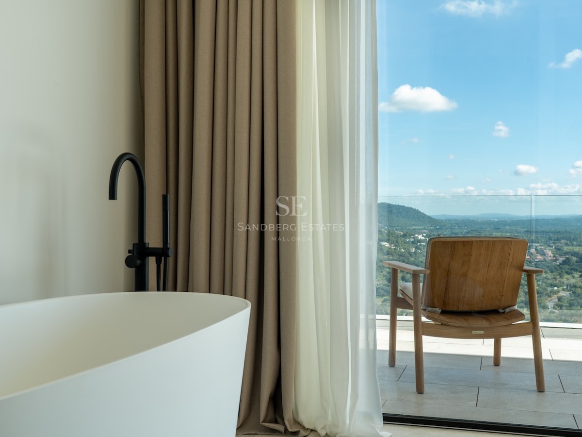 White freestanding bathtub and black faucet next to a floor-to-ceiling glass door opening onto a terrace with views.