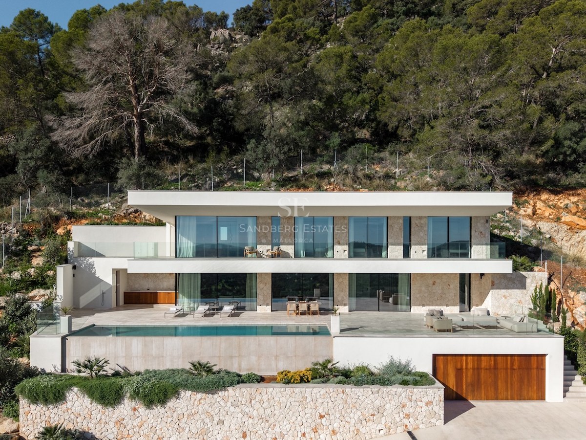 Front view of a two-story modern white villa with large glass windows, a swimming pool, and stone walls set against a forest.