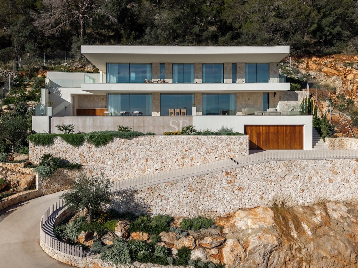 Modern white villa built into a rocky hillside with dry stone walls, large floor-to-ceiling windows, and a curved driveway.