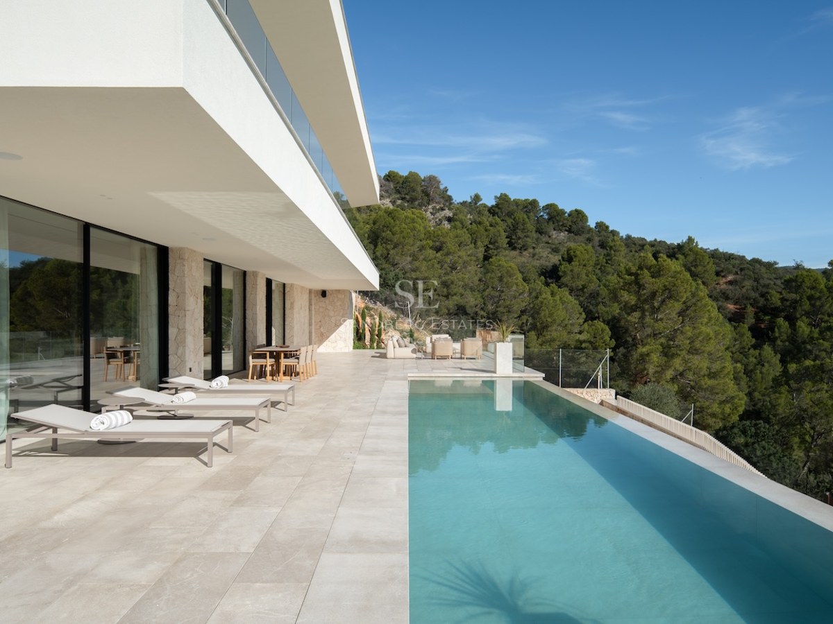 A spacious white stone terrace featuring an infinity pool, sun loungers, and views of a pine forest under a blue sky.
