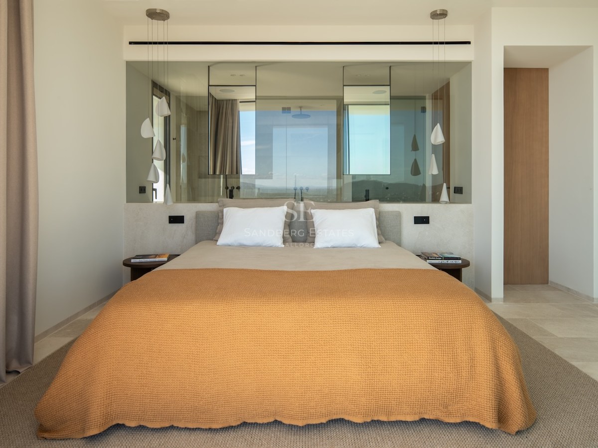 Modern bedroom featuring an orange bedspread, glass partition wall behind the bed, and sculptural white pendant lamps.