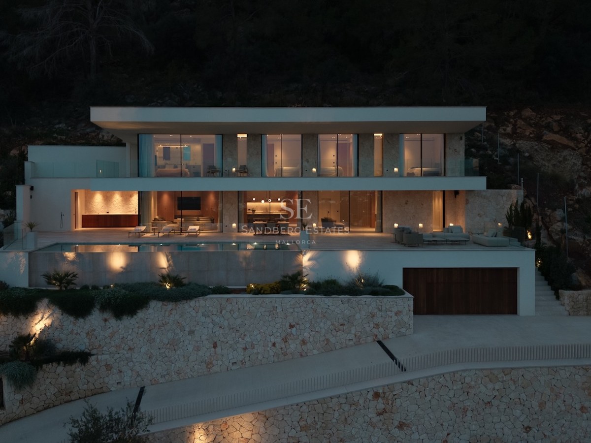 Illuminated nighttime facade of a three-level modern villa featuring an infinity pool and natural stone walls.