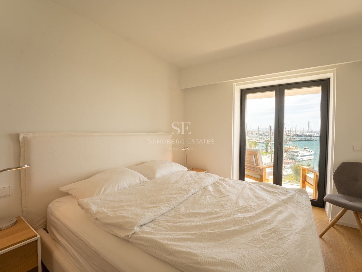 Chambre lumineuse avec lit blanc, chaise grise et baies vitrées donnant sur un port de plaisance.