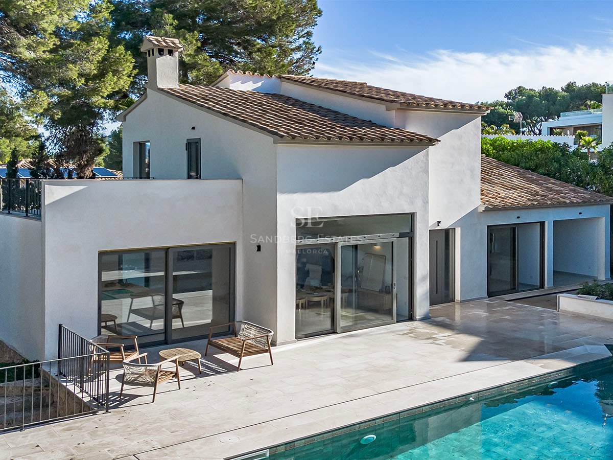 Villa blanca moderna con techo de tejas, puertas de cristal y terraza de piedra junto a una piscina turquesa.