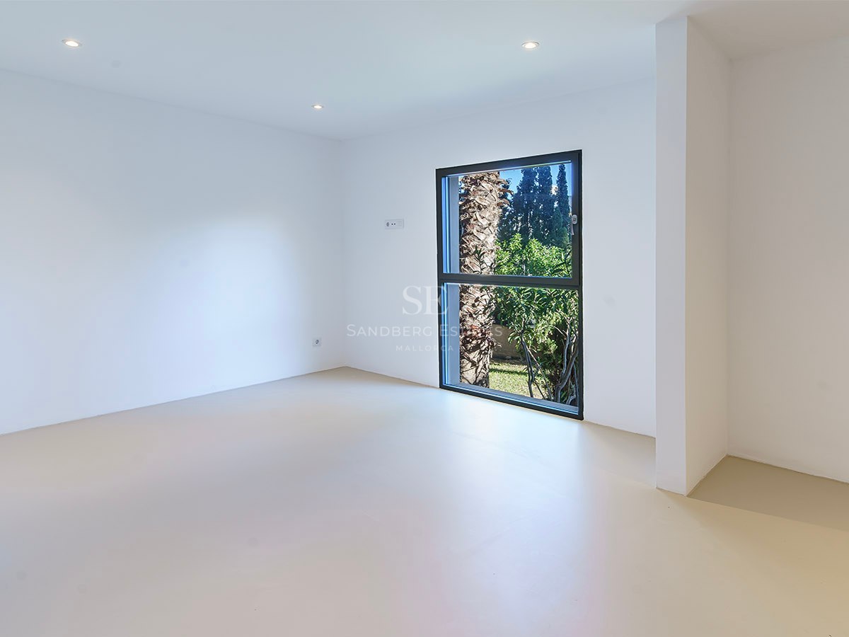 Habitación minimalista vacía con paredes blancas, suelo claro y ventana vertical con vistas a una palmera.
