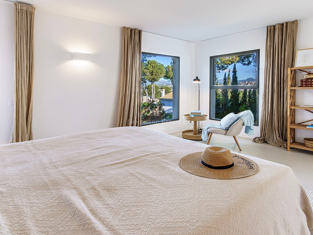 Dormitorio moderno con paredes blancas, cortinas de lino, muebles de madera y vistas a un paisaje verde.
