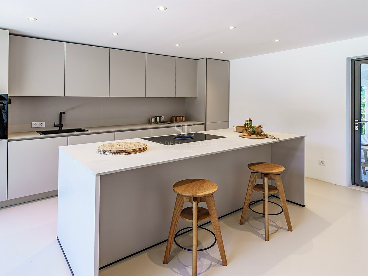 Elegante cocina gris claro con una gran isla, taburetes de madera y puerta de cristal hacia la terraza.