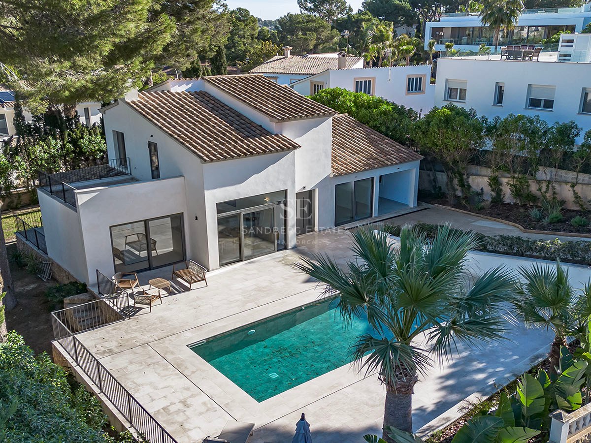 Vista aérea de una villa blanca con tejado de teja, piscina turquesa y amplia terraza rodeada de pinos y palmeras.