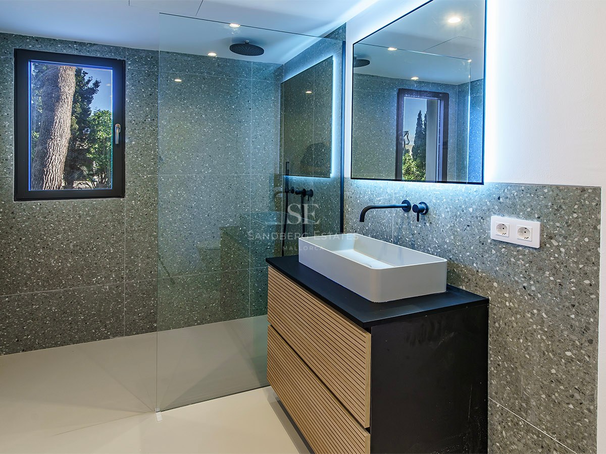 Baño moderno con paredes de terrazo gris, ducha de cristal, grifería negra y mueble de madera con lavabo blanco.