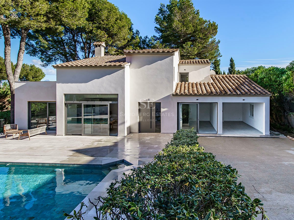 Villa blanca moderna con piscina turquesa, terraza de piedra y pinos bajo un cielo azul despejado.