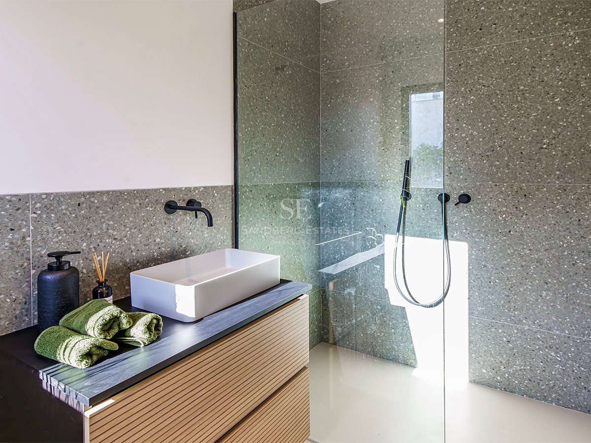 Baño moderno con azulejos grises moteados, lavabo blanco sobre mueble de madera y ducha con mampara de cristal.
