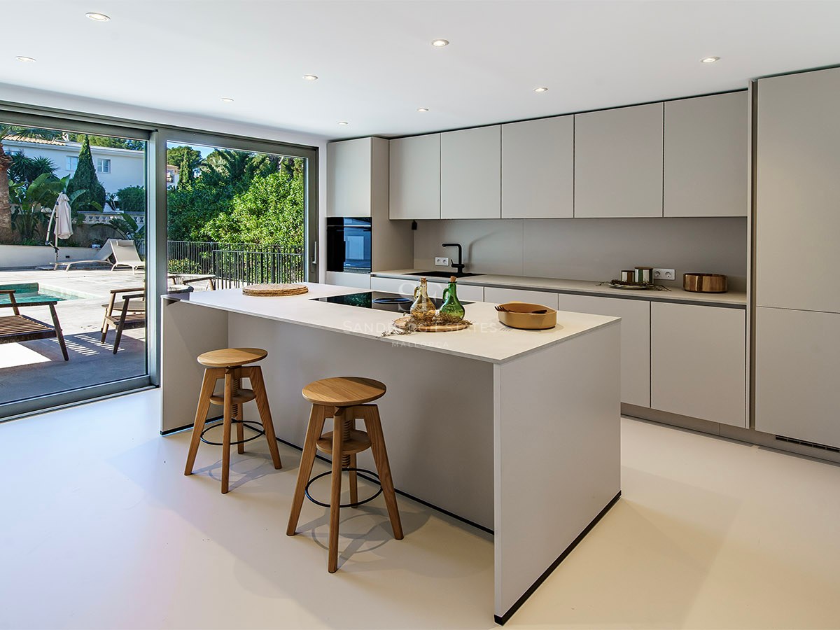 Cocina moderna blanca con isla central, dos taburetes de madera y grandes puertas correderas de cristal hacia la piscina.