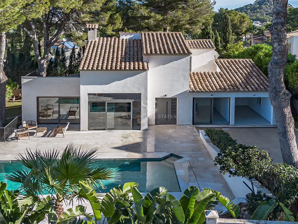 Vista aérea de una villa blanca con techo de teja, piscina turquesa y terraza de piedra rodeada de pinos.
