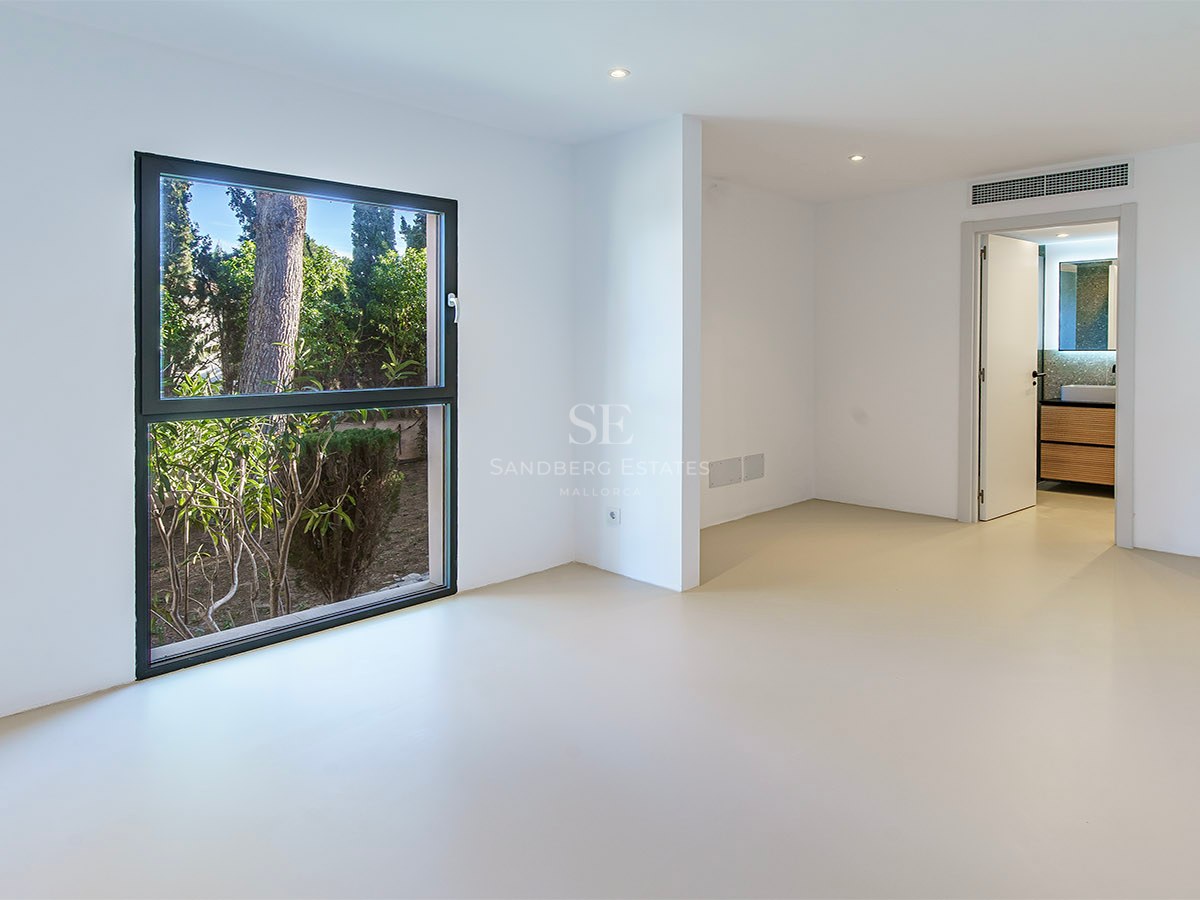 Dormitorio minimalista vacío con paredes blancas, suelo beige, ventana al jardín y puerta a un baño en suite.