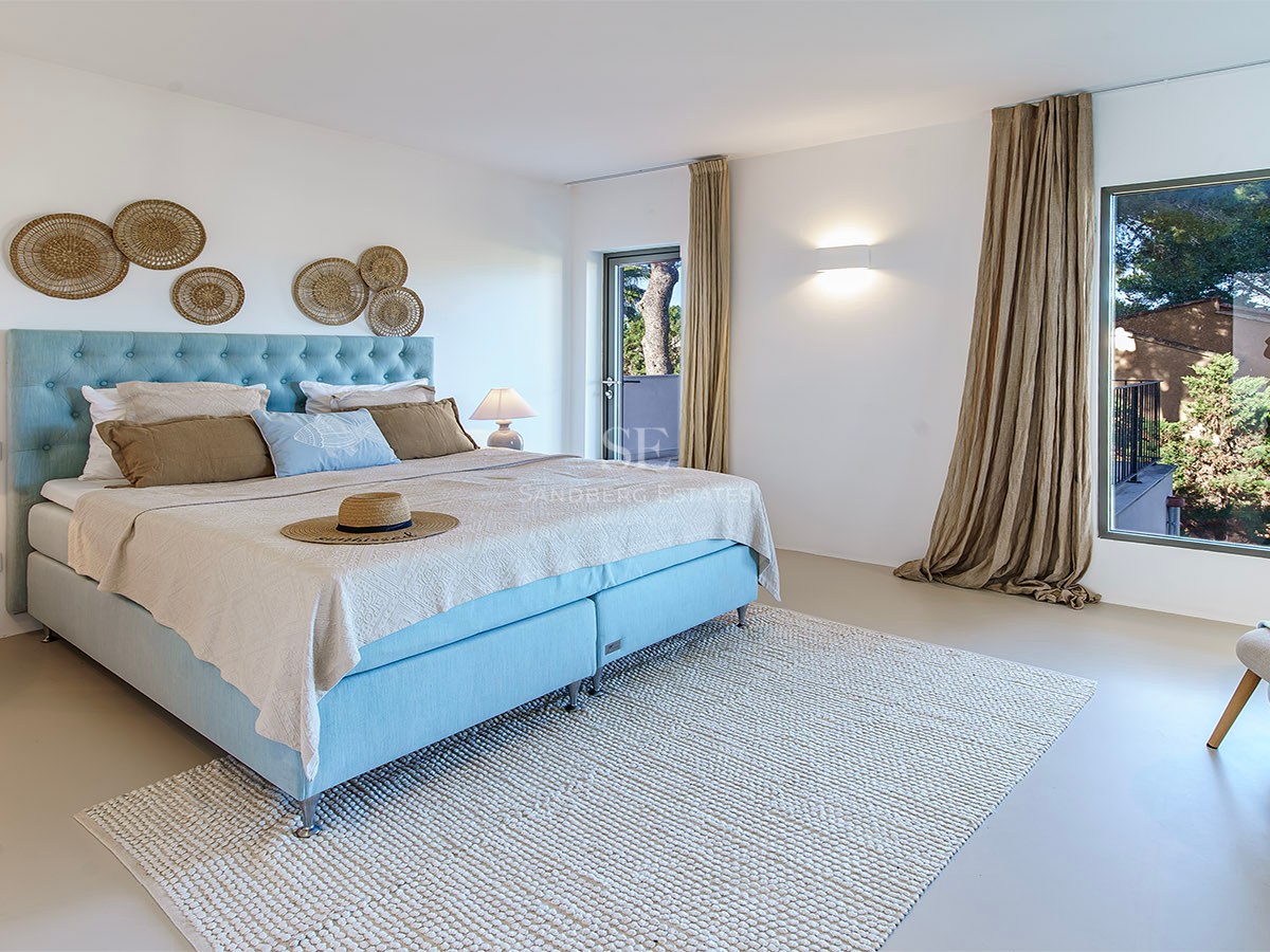 Habitación luminosa con cama azul claro, alfombra de lana y gran ventanal con vistas a árboles verdes.