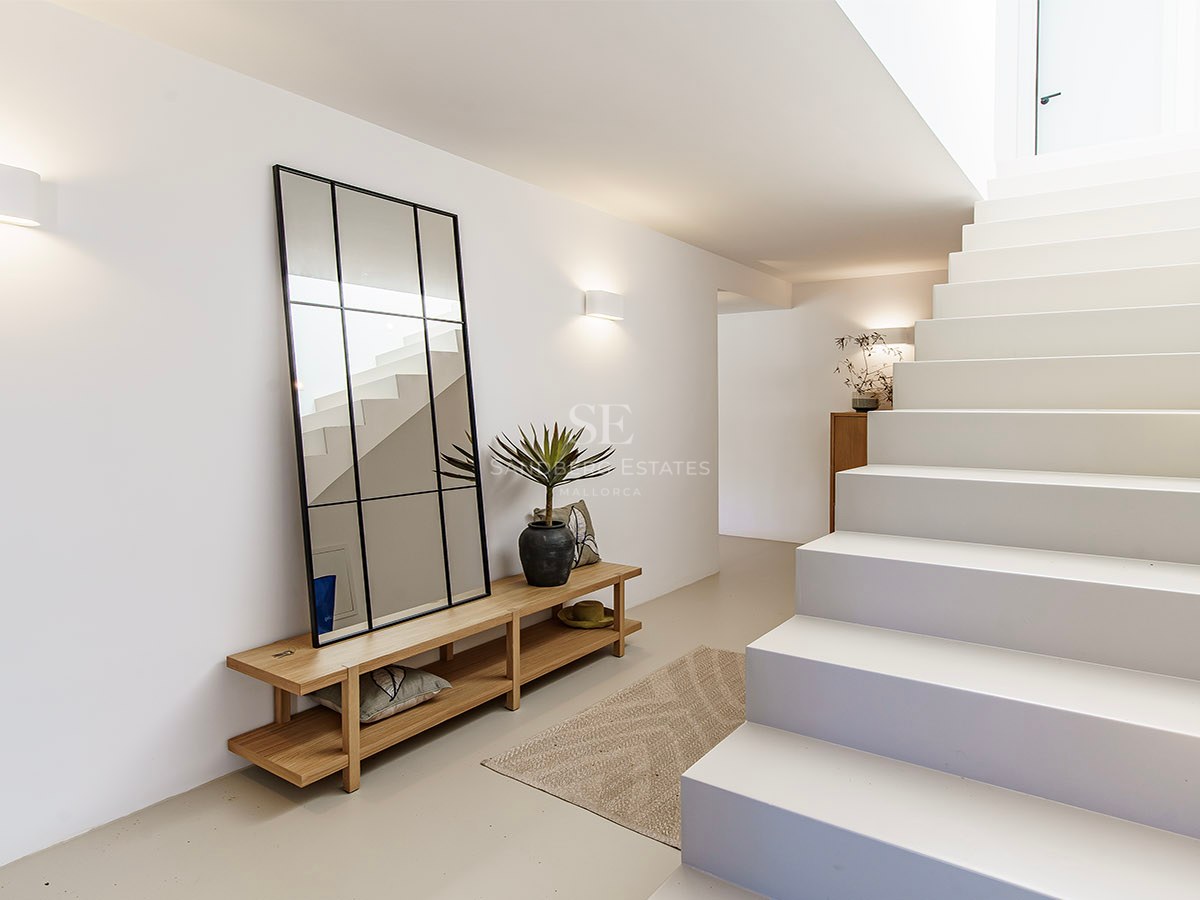 Pasillo minimalista luminoso con escaleras blancas, un gran espejo de marco negro, banco de madera e iluminación cálida.