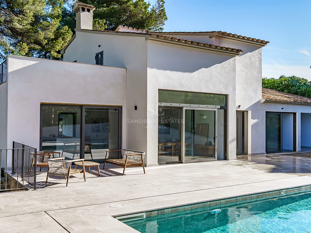 Una gran piscina turquesa frente a una villa blanca moderna con terraza de piedra y muebles de exterior.