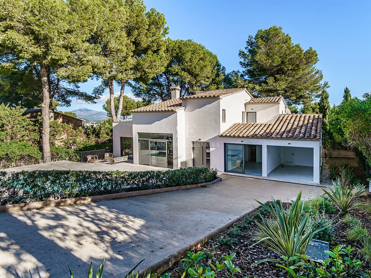 Una villa blanca moderna de dos plantas con tejas de terracota, rodeada de altos pinos y una zona de piscina.