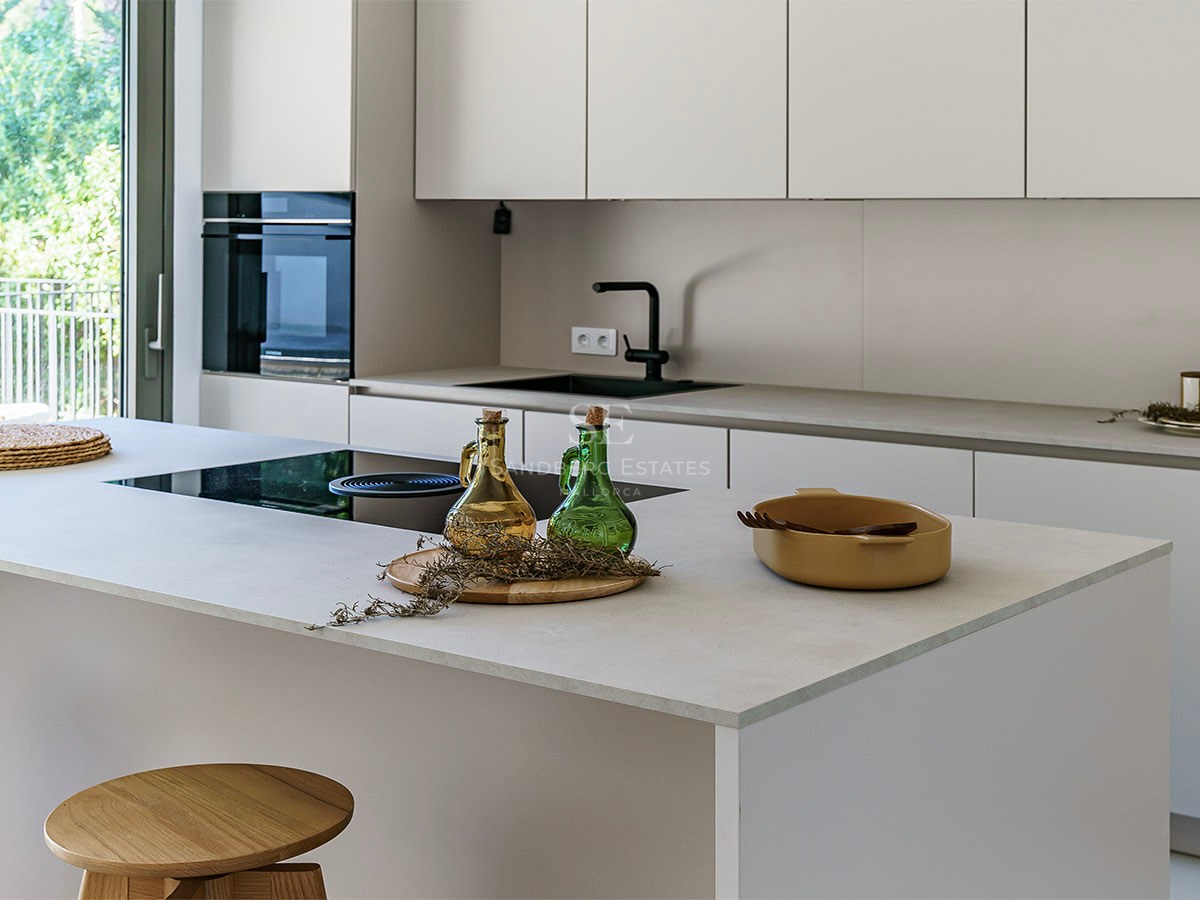 Cocina blanca minimalista con isla central, taburetes de madera y ventanal con vistas a un jardín frondoso.