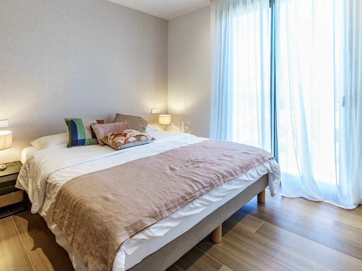 Chambre lumineuse avec un grand lit, des coussins décoratifs, des lampes de chevet et de grandes fenêtres avec voilages.