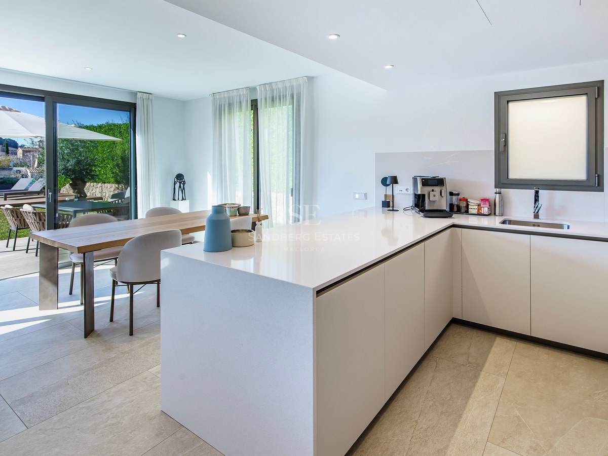 Cuisine minimaliste blanche avec îlot central, table à manger en bois et baies vitrées ouvrant sur un jardin.