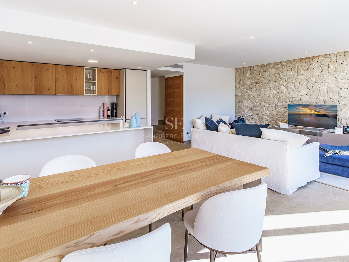 Pièce lumineuse à aire ouverte avec table à manger en bois, cuisine moderne, canapé blanc et mur en pierre naturelle.