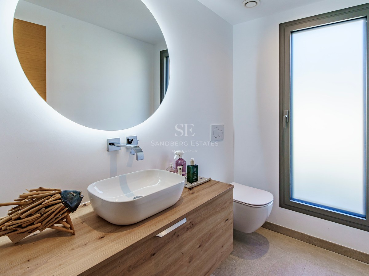 Salle de bain moderne avec vasque sur meuble en bois, miroir rond éclairé et fenêtre en verre dépoli.