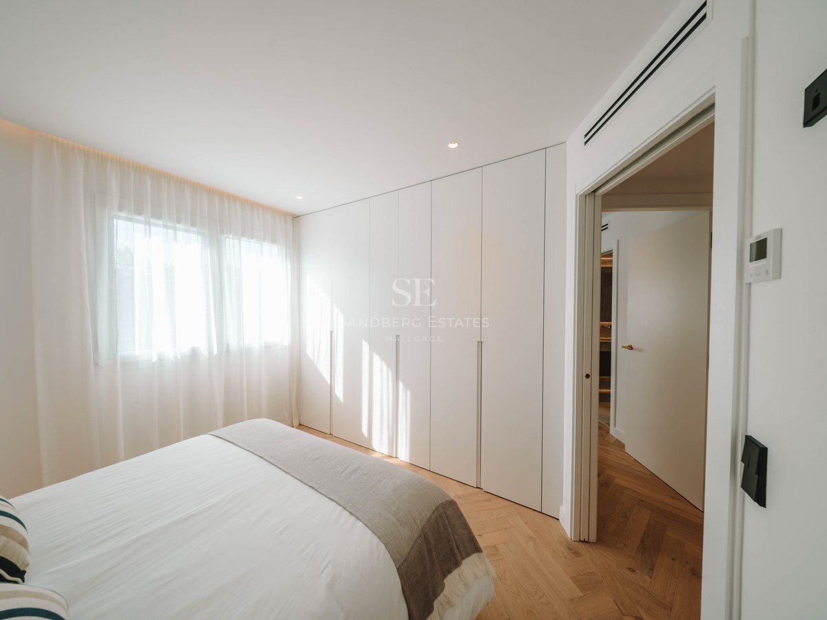 Bright bedroom featuring herringbone wood floors, floor-to-ceiling white wardrobes, a bed with white linens, and sheer curtains.