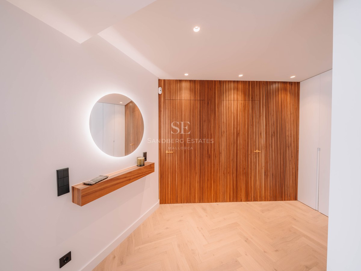 Minimalist hallway featuring a backlit circular mirror, wooden floating shelf, and vertical wood slat wall.